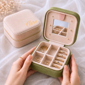 Puede incluir: Un joyero cuadrado de terciopelo verde oliva con interior beige, que incluye un espejo y múltiples compartimentos para anillos y collares. Un joyero a juego de terciopelo beige con el nombre Clara impreso en dorado se encuentra al lado.