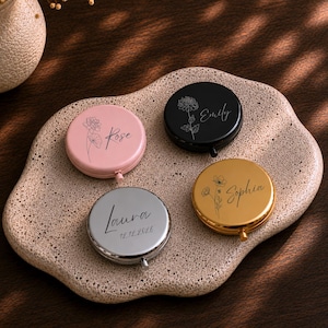 Puede incluir: Cuatro espejos de bolsillo redondos con acabados en rosa, negro, dorado y plateado. Cada espejo presenta un grabado floral de líneas único y un nombre personalizado. El espejo plateado incluye la fecha 12.12.2026 grabada bajo el nombre Laura.