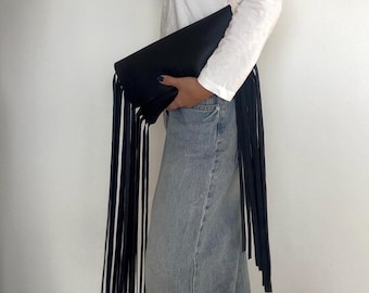 Peut inclure: Une pochette noire dotée de longues franges fines qui pendent sur les côtés. Le sac présente une surface noire lisse et unie, tenue horizontalement pour mettre en valeur la longueur des brins de franges sur un fond uni et neutre.