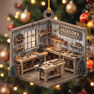May include: A wooden ornament shaped like a workshop interior with the text "JACKSON'S WORKSHOP" on the wall. The scene features multiple workbenches, various woodworking tools, shelves, a window, and two small decorative pine trees.