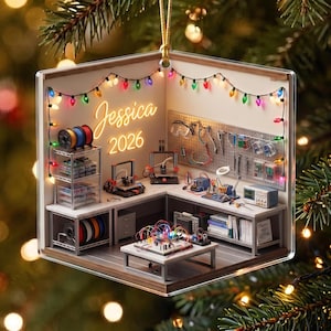 May include: A square Christmas ornament featuring a 3D miniature workshop scene with a pegboard, 3D printers, and electronic components. The walls are decorated with colorful string lights and the text Jessica 2026 in glowing orange script.