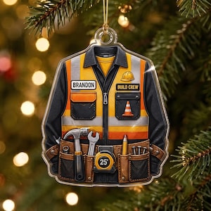 May include: A clear acrylic ornament shaped like a work shirt with high-visibility orange and black panels. It features a name tag reading BRANDON, a BUILD CREW patch, a hammer, wrench, tape measure labeled 25', pencil, and nails in pockets.