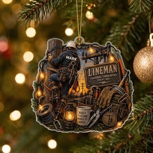 May include: A decorative ornament featuring a lineman climbing a utility pole with the name MARK on the back of the jacket. The design includes a sign reading LINEMAN POWERING COMMUNITIES EVERY DAY and a mug labeled BUILT TO CLIMB WIRED TO SERVE.