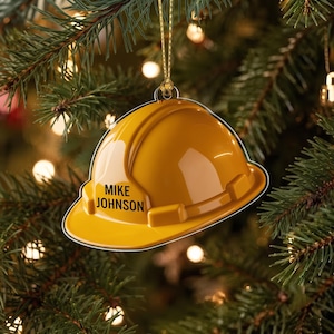 May include: A yellow hard hat ornament featuring the name MIKE JOHNSON printed in black capital letters on the front. The ornament is suspended by a thin gold cord and has a smooth, glossy finish with a realistic construction helmet shape.