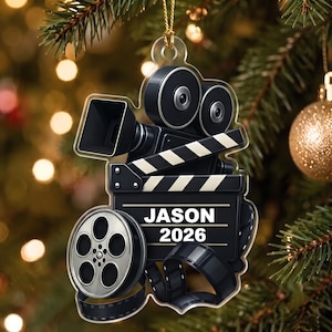 May include: A flat, transparent acrylic ornament shaped like a movie camera, film reel, and clapperboard. The black clapperboard features the white text JASON and 2026. The design includes a silver film reel and a black camera lens with film strips.