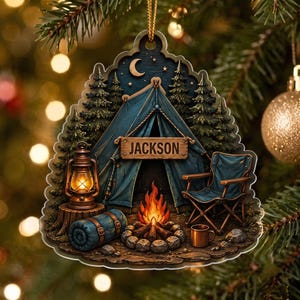 May include: A decorative ornament featuring a camping scene with a blue tent, a campfire, a lantern, a sleeping bag, and a folding chair. A wooden sign hanging on the tent displays the name JACKSON. The background shows pine trees and a crescent moon.