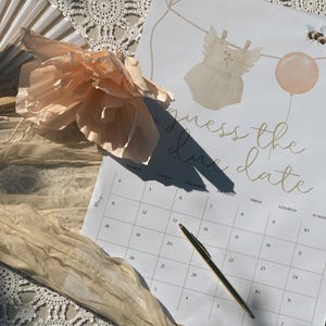 Könnte beinhalten: Ein weißer Papierkalender mit dem Text Guess the due date, verziert mit Illustrationen eines Baby-Stramplers, eines Luftballons und einer Biene. Ein goldener Stift liegt auf dem Kalenderraster. Eine pfirsichfarbene Papierblume und ein Fächer liegen daneben.