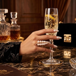 Puede incluir: Un anillo llamativo de tono dorado con forma de hoja, que presenta múltiples perlas blancas pequeñas dispuestas a lo largo de las nervaduras. El anillo se lleva en un dedo mientras se sostiene una copa de champán de cristal con líquido dorado.