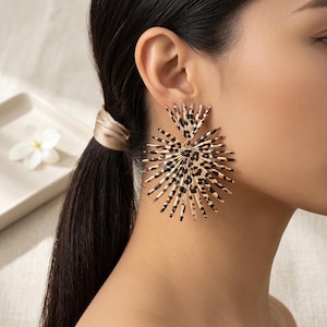 Può includere: Un grande orecchino circolare con un design a raggiera composto da sottili punte metalliche radianti. La superficie dell'orecchino è rifinita con un motivo leopardato nei toni del beige e del nero, creando un look audace e ad alto contrasto.