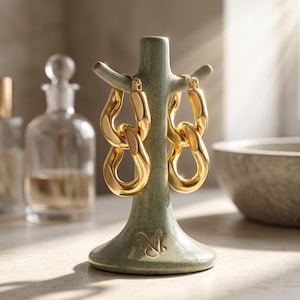 Puede incluir: Un par de pendientes de aro dorados con un diseño de eslabones de cadena gruesos y entrelazados, expuestos en un soporte de cerámica verde salvia con forma de pequeño árbol, base acampanada y un logotipo monograma en relieve.