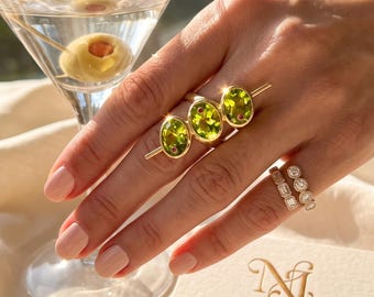 Peut inclure: Une bague en or ornée de trois péridots vert lime taille ovale avec de petits accents rouges, portée avec deux alliances en diamant superposées. Une carte de visite avec le texte Nesa Jewelry & Co. est visible, ainsi qu'un verre à martini.