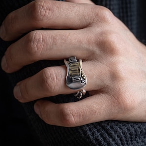 May include: A silver-toned ring featuring a miniature electric guitar design with a gold-coloured pickguard and fretboard detail. The ring has an open, adjustable band with a small beaded texture at the ends, worn on a finger.