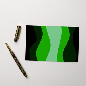 May include: A rectangular card featuring a vertical wavy pattern in shades of black, dark green, bright green, and light mint green. Beside the card lies a black ballpoint pen with a gold floral pattern and a matching removable pen cap.