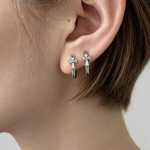 May include: A pair of silver-toned hoop earrings, each featuring a small, detailed cross charm attached to the front. The earrings have a polished metallic finish and a hinged closure mechanism, designed to sit securely on the earlobe.