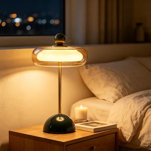 May include: A table lamp featuring a dark green dome base, a slender gold-toned metal stem, and a wide, transparent amber glass shade with a central white diffuser. A small dark green sphere sits atop the center of the glass shade.