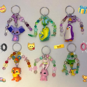 May include: Six keychains featuring various plastic animal figures and colourful beads. The top row includes a purple koala, a yellow dolphin, and a purple penguin. The bottom row features an orange lion, a pink rabbit with a cake, and a green creature.