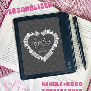 May include: A black Kobo e-reader displaying a dark gray screensaver with a white heart-shaped arrangement of books and the text Angela's LIBRARY. A black stylus rests on the right side of the device, which is placed on an open book.