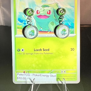 May include: A pair of dangle earrings featuring a small green leaf symbol stud connected by a silver chain to a larger circular charm displaying a Bulbasaur character. The earrings are mounted on a green Bulbasaur trading card with text.