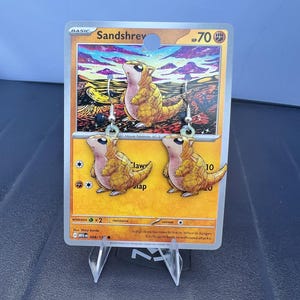 May include: A pair of dangle earrings featuring the yellow, armoured Sandshrew character from the Pokémon franchise. The earrings are displayed on a Sandshrew trading card, which shows the character in a desert landscape with a purple and orange sky.