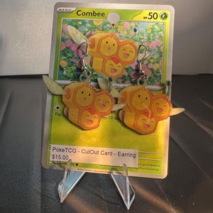 Puede incluir: Un par de pendientes colgantes con el Pokémon Combee amarillo en forma de panal, presentados sobre una carta coleccionable Pokémon verde. Los pendientes están hechos de cartulina recortada y cuelgan de ganchos de metal plateado con tres caras sonrientes.