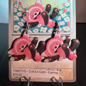 May include: A pair of dangle earrings featuring the pink and brown Stufful Pokémon character with a glittery finish. The earrings are displayed on a Stufful trading card that shows the character in a field of flowers with the text Stufful and HP 70.