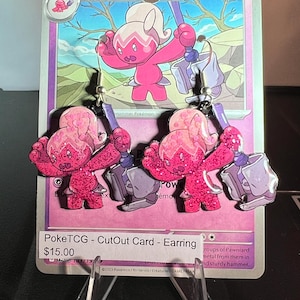 Puede incluir: Un par de pendientes colgantes con el Pokémon rosa Tinkatuff sosteniendo un gran martillo gris. Los pendientes son brillantes y están suspendidos de ganchos plateados. Se muestran sobre una carta coleccionable Pokémon morada con el texto PokeTCG - CutOut Card - Earring.