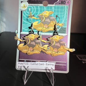 May include: A pair of dangling earrings featuring the yellow Kadabra character holding a silver spoon, attached to a purple Kadabra trading card display backing. The card displays the text Kadabra, HP 80, and PokeTCG - CutOut Card - Earring.
