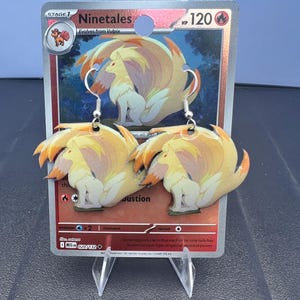 May include: A pair of dangle earrings featuring the Ninetales character from a Pokemon trading card. The earrings are shaped like the nine-tailed fox with yellow and orange fur, suspended from silver-toned metal hooks against the original card.