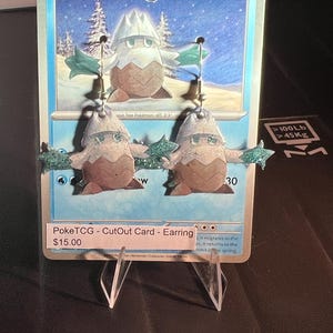 May include: A pair of dangle earrings featuring the character Snover, crafted from a cutout of a blue Pokémon trading card. The earrings display the brown and white creature with green arms, suspended by silver-toned metal hooks against the original card.