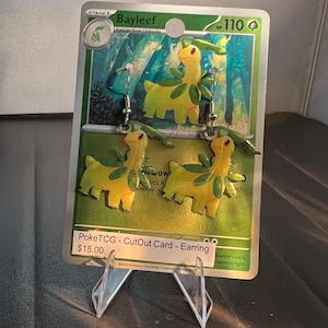 May include: A pair of dangle earrings shaped like the yellow and green creature Bayleef, suspended from silver-toned metal hooks. The earrings are displayed on a green Pokémon trading card backing that features the same character illustration.