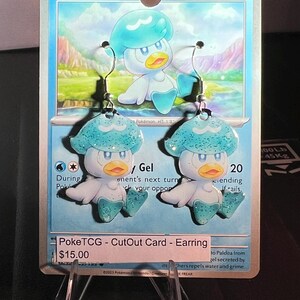 Puede incluir: Un par de pendientes colgantes con el personaje Quaxly, hechos a partir de recortes de una carta coleccionable Pokémon azul. Los pendientes muestran al personaje con un gorro azul verdoso, pico amarillo y pies azules en ganchos plateados.