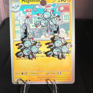 May include: A pair of dangle earrings featuring the Magneton character from the Pokémon trading card game. The earrings are shaped like the light blue, three-part robotic creature with red and blue magnets, suspended from silver-toned metal hooks.