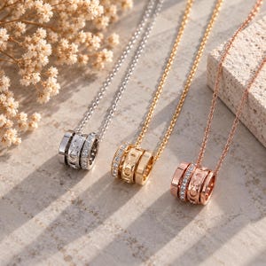 Può includere: Tre collane con pendenti cilindrici nei toni dell'argento, dell'oro e dell'oro rosa. Ogni pendente presenta una fascia centrale con il logo Coach e due fasce esterne incastonate con piccoli cristalli chiari, su catene metalliche sottili.