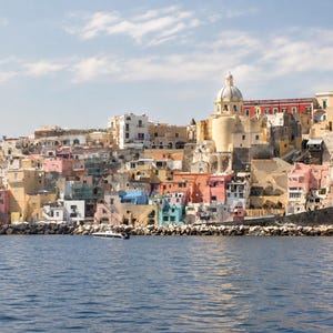 May include: A coastal townscape featuring a dense cluster of multi-colored buildings in shades of yellow, pink, and white, dominated by a large church with a central dome, situated along a rocky shoreline overlooking a deep blue body of water.
