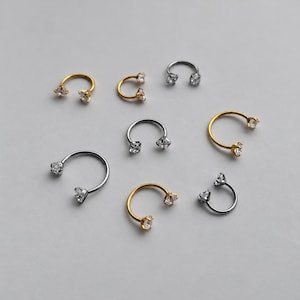 May include: A collection of eight horseshoe-shaped body jewelry pieces in gold and silver tones. Each circular barbell features a clear, faceted gemstone set at both ends. The jewelry varies in size and metal finish, displaying a metallic luster.