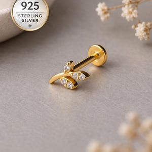 Könnte beinhalten: Ein goldfarbener Ohrstecker mit flacher Rückseite und einem zweigartigen Design mit drei kleinen Blättern, die jeweils mit einem klaren, funkelnden Stein besetzt sind. Das Schmuckstück ist als 925 Sterling Silber gekennzeichnet.