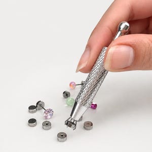 May include: A silver-toned metal prong tool with a textured grip handle is shown holding a small circular stud earring. Several other stud earrings with various colored stones and flat metallic tops are scattered around the tool.