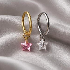 Könnte beinhalten: Zwei Creolen mit baumelnden sternförmigen Anhängern. Ein Ring ist goldfarben mit einem durchscheinenden rosa Stern, der andere ist silberfarben mit einem klaren, facettierten Stern. Beide Ringe haben eine glatte, polierte Metalloberfläche.