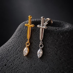 Op de afbeelding: Twee zwaardvormige lichaamsieraden, één in goudkleur en één in zilverkleur, elk met een verticaal gevest- en lemmetontwerp met een kleine, heldere, marquise-geslepen edelsteen die met een ringetje aan de punt is bevestigd.