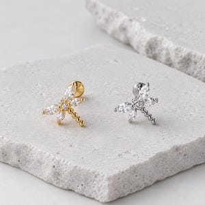 Può includere: Due orecchini a lobo a forma di libellula, uno in metallo dorato e uno in metallo argentato. Ogni orecchino presenta cristalli trasparenti taglio marquise che formano le ali e una coda in metallo perlato, con una chiusura circolare piatta.