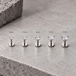 Pode incluir: Cinco brincos labret prateados com gemas transparentes de corte redondo, dispostos em fila do menor para o maior. Os tamanhos estão indicados acima de cada peça como 2mm, 2,5mm, 3mm, 4mm e 5mm de diâmetro.
