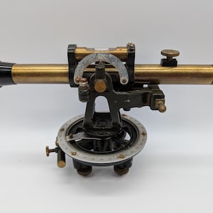 May include: A vintage brass and black metal surveyor's level instrument featuring a horizontal telescope, a spirit level vial, a semi-circular vertical arc, and a circular base plate with engraved degree markings and adjustment knobs for precise alignment.