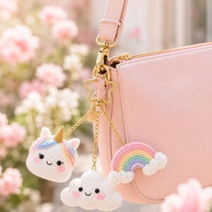 Puede incluir: Un bolso de mano rosa claro con un cierre dorado en forma de estrella del que cuelgan tres dijes de ganchillo: un unicornio blanco con cuerno dorado y crin pastel, una nube blanca con mejillas rosadas y un arcoíris colorido.