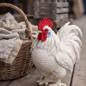 May include: A handcrafted crochet rooster figure featuring a white body with textured stitch details, a bright red comb and wattle, and a small blue ear patch. The rooster has dark eyes, a light-colored beak, and detailed plumage on its tail.
