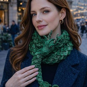 May include: A green knitted scarf shaped like a dragon with textured scale-like patterns, a detailed head with small horns, and a curled tail. The scarf is wrapped around the neck, featuring deep emerald and lighter green yarn tones throughout.