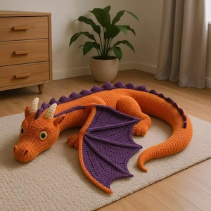 Op de afbeelding: Een groot, gehaakt draak-speeltje met een feloranje lichaam en een lange, gebogen staart. Het heeft paarse vleugels met gedetailleerde stiksels, een rij paarse stekels langs de rug en twee kleine, crèmekleurige hoorns op de kop met grote ogen.
