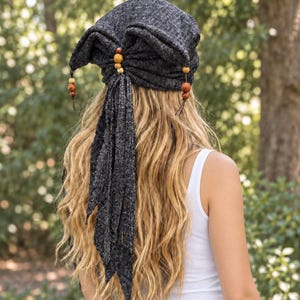 May include: A dark charcoal grey knit head wrap featuring a gathered back design with wooden beads and long, dangling fabric ties. The textured fabric has a subtle heathered appearance, and the accessory is secured with a central bead accent.