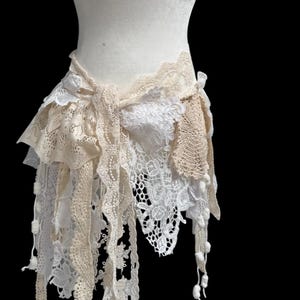 Puede incluir: Un cinturón de encaje crema y blanco, exhibido en un maniquí. El cinturón presenta múltiples capas de encaje en diversas texturas y patrones, con un lazo anudado en la parte delantera. El diseño incluye encaje colgante y pequeñas bolitas decorativas.