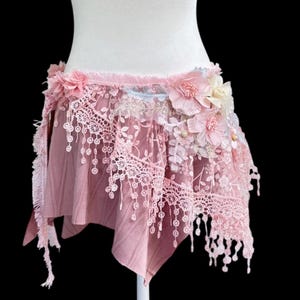 May include: Pink, layered skirt with lace trim and floral accents. The skirt is displayed on a white mannequin against a black background. The skirt has a fringed hem and is decorated with pink and white flowers.