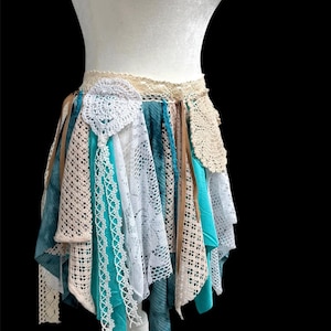 Puede incluir: Falda bohemia exhibida en un maniquí. Presenta telas turquesas, blancas y beige, incluyendo encaje y cintas. La falda tiene un dobladillo asimétrico y una cintura de ganchillo.